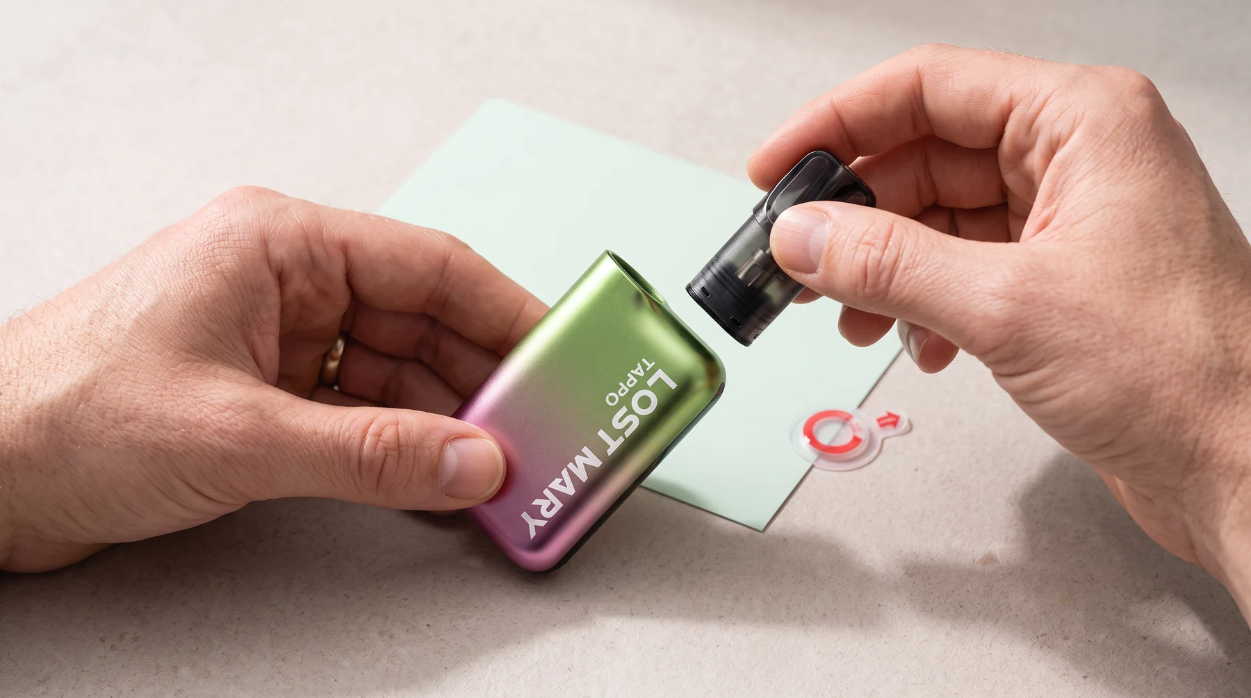 Adult hands aligning a black Lost Mary Tappo pod above the green-and-pink battery device after removing its protective seal